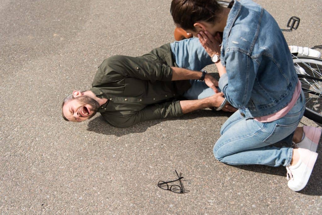 A man lies on the ground in pain with an injured leg near a bicycle, while a woman kneels beside him, appearing concerned. A pair of glasses is on the ground nearby.
