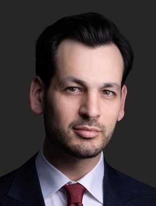 Person with dark hair, light skin, wearing a dark suit, white shirt, and red tie, against a dark background.