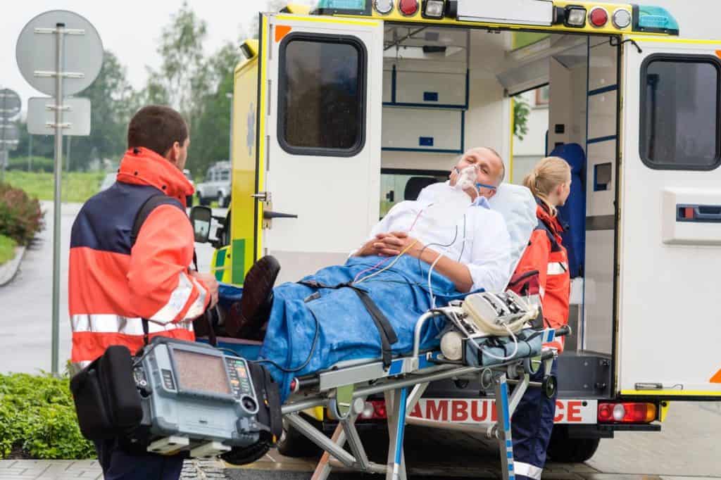 Paramedics load a patient on a stretcher into an ambulance. The patient is wearing an oxygen mask. Medical equipment is visible next to the stretcher.