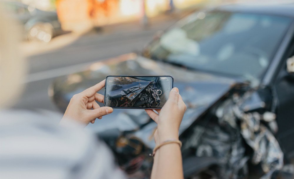 Person photographing the aftermath of a car accident with a smartphone, showing damage to the vehicle's front end.