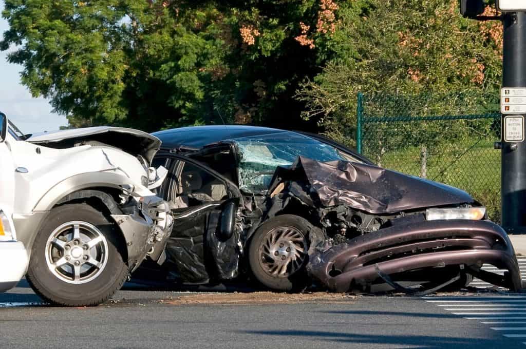 A white vehicle and a dark-colored car are involved in a serious collision at an intersection, with extensive damage to both front ends. Trees and a fence are visible in the background.